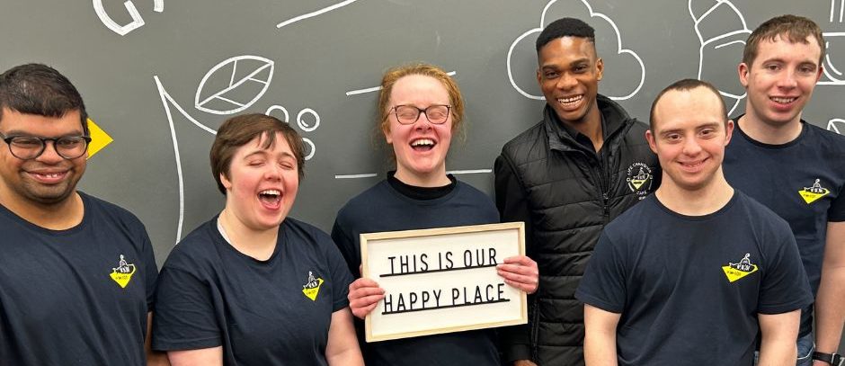 Group of trainees wearing grey t-shirts smiling whilst holding a sign that says